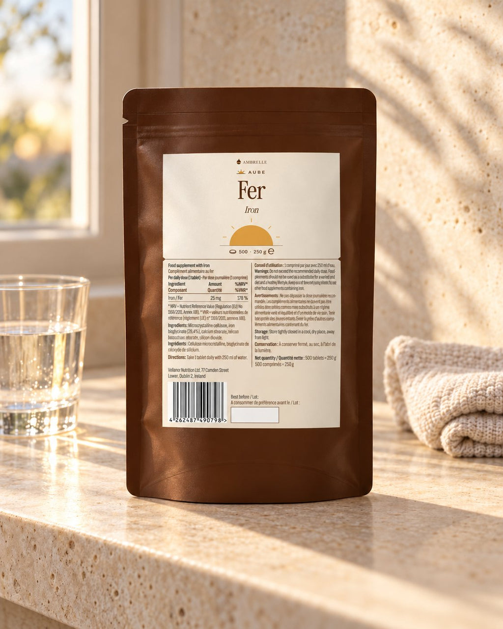 Iron Bisglycinate 25 mg by AMBRELLE on a travertine bathroom shelf at sunrise, with a glass of water