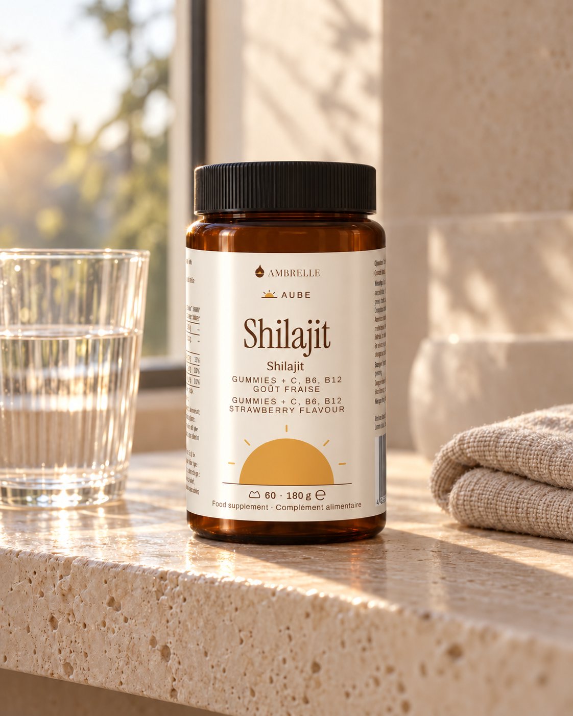 Shilajit Gummies + C, B6, B12 by AMBRELLE on a travertine bathroom shelf at sunrise, with a glass of water